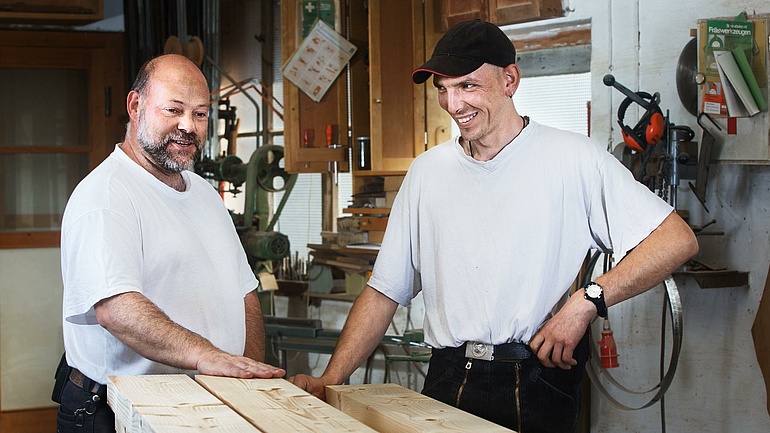 Zwei lächelnde Männer in weißen Shirts in einer Holzwerkstatt.