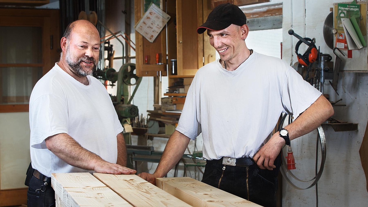 Zwei lächelnde Männer in weißen Shirts in einer Holzwerkstatt.