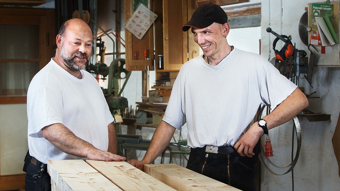 Zwei lächelnde Männer in weißen Shirts in einer Holzwerkstatt.